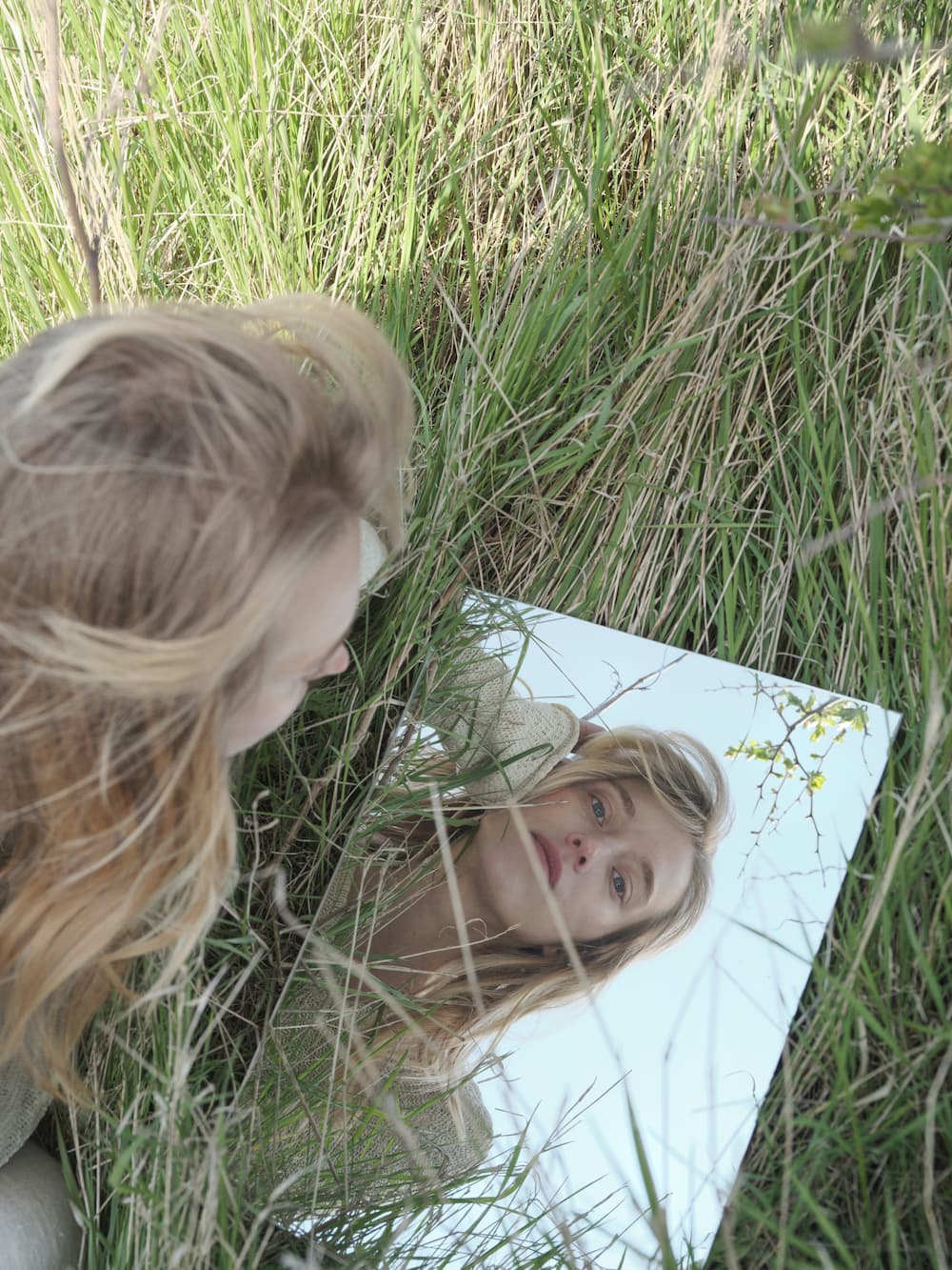 Woman looking at her reflection in a mirror surrounded by tall grass