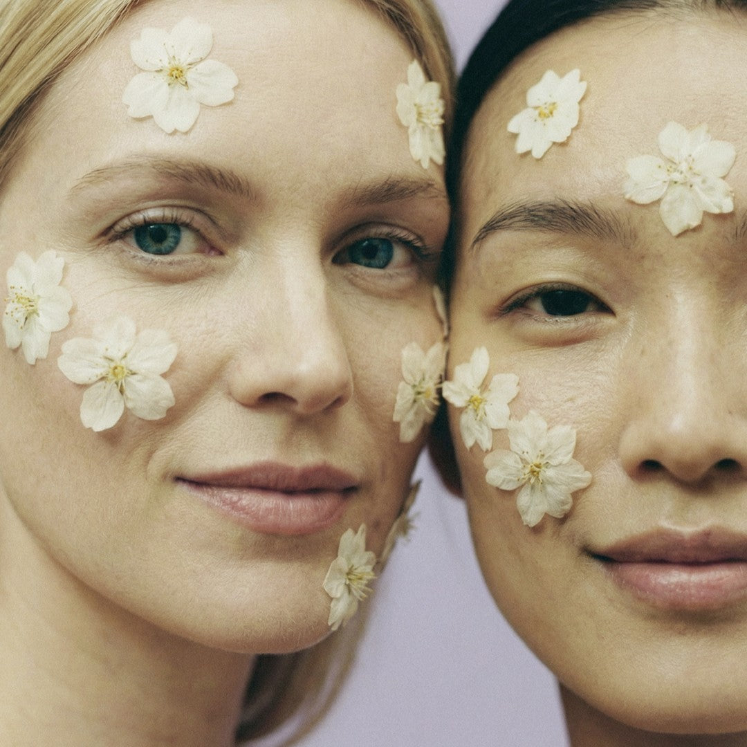 Two women with smooth skin and white flower petals on their faces, symbolizing natural beauty and skin longevity.