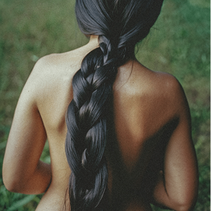 Woman with healthy, glowing skin and a long thick braid running down her bare back against a natural green background.