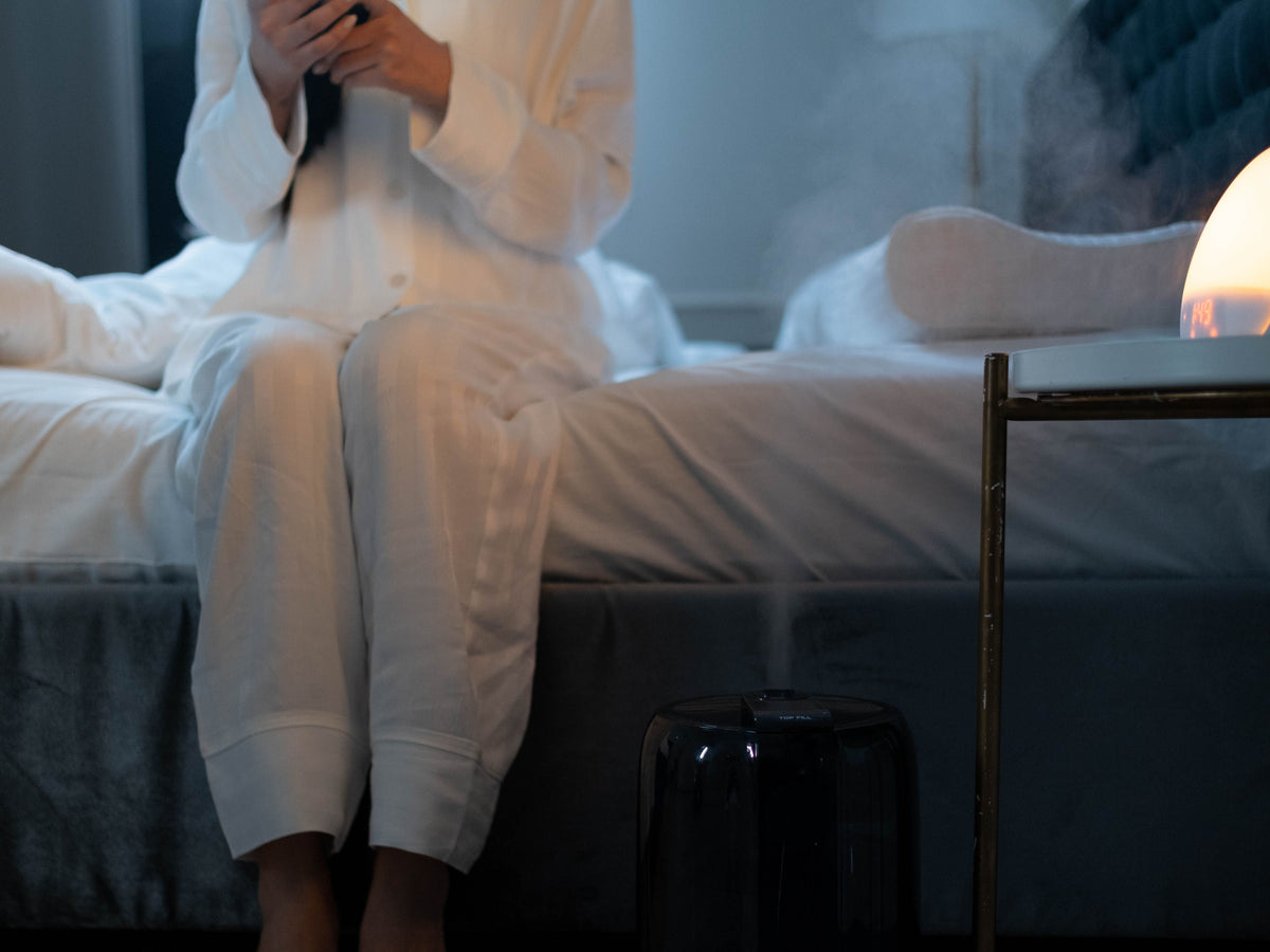 A person in white pajamas sits on a bed in a dimly lit bedroom, holding a phone. A small black diffuser is visible in the foreground, with a glowing lamp on a side table to the right.